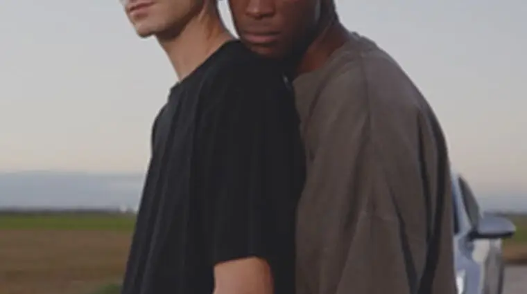 A gay couple standing under the Motto logo.